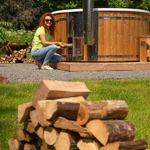 Outdoor-Foto einer rothaarigen Frau, die neben einem holzbeheizten Badefass mit Ofen auf einem Schotterpodest hockt; im Vordergrund unscharf ein aufgestapeltes Brennholz.