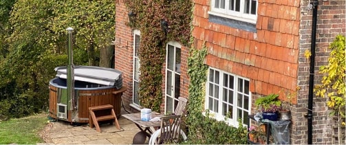 Outdoor view of wood fired hot tub with stainless steel stove and wooden steps beside brick house with ivy and garden plants
