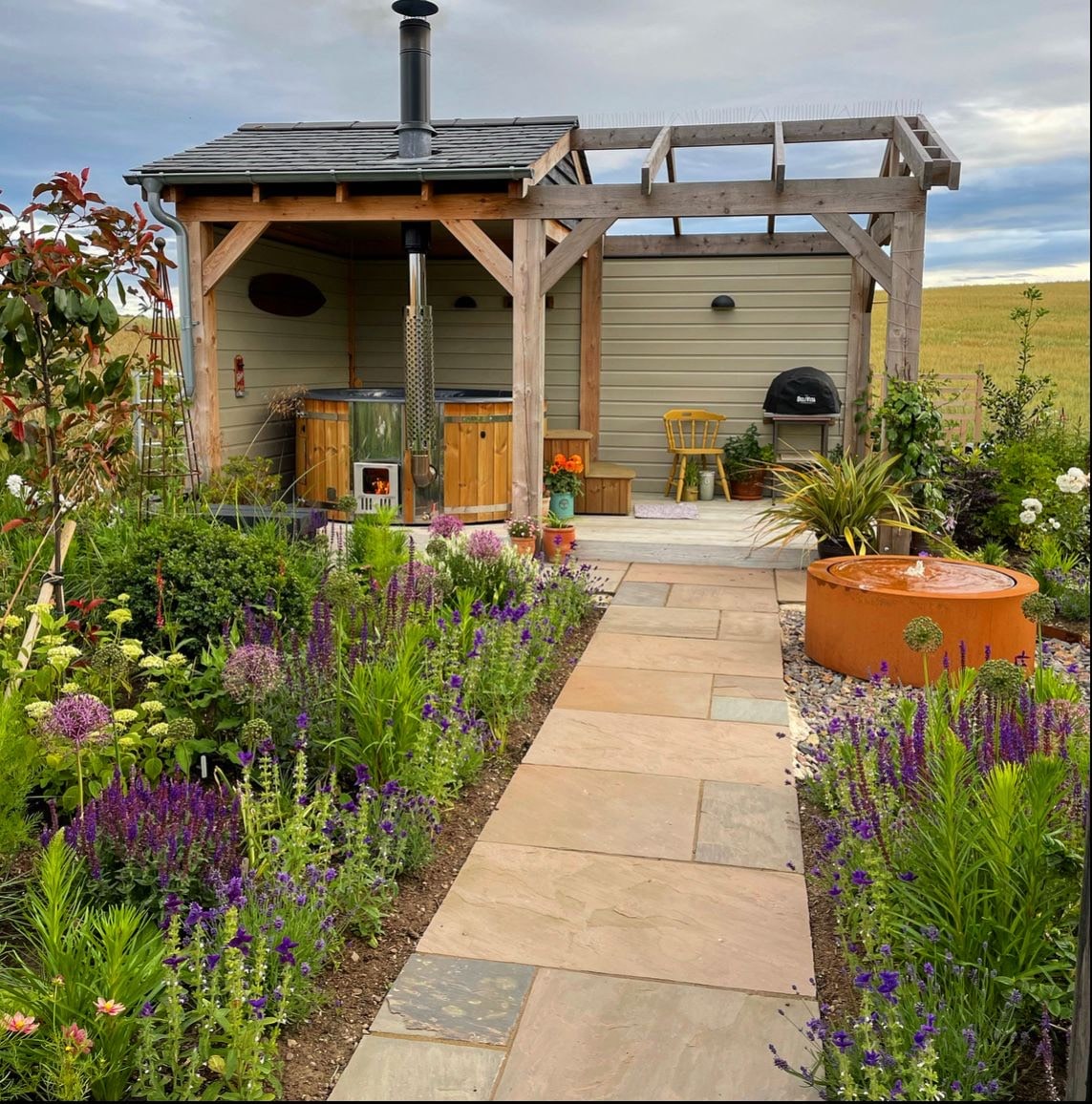 Gardenvity wood fired hot tub in countryside garden with wooden pergola and flowers.