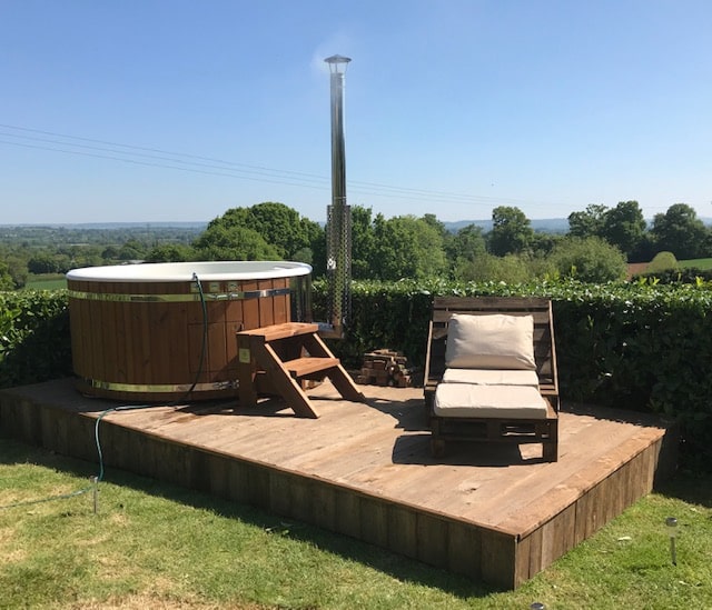 Gardenvity wood fired hot tub on raised wooden deck with countryside view and sun loungers.